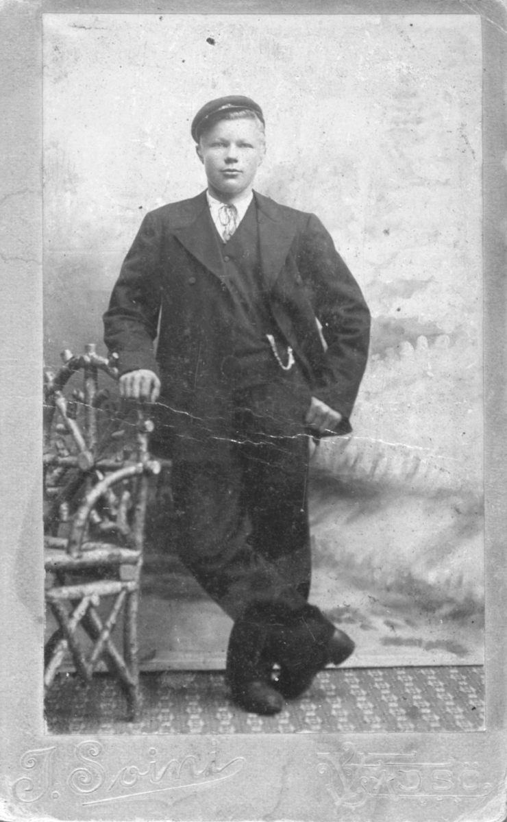 En ung gutt lener seg til en stol på et fotostudio. Han er kledd i dress: jakke, vest, bukser og et lommeur. Han har også på seg en hatt. Portrettet muligens tatt på slutten av 1800-tallet eller begynnelsen av 1900-tallet.