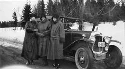 Tre kåpekledte damer foran en bil. Snø og grantrær i bakgrun