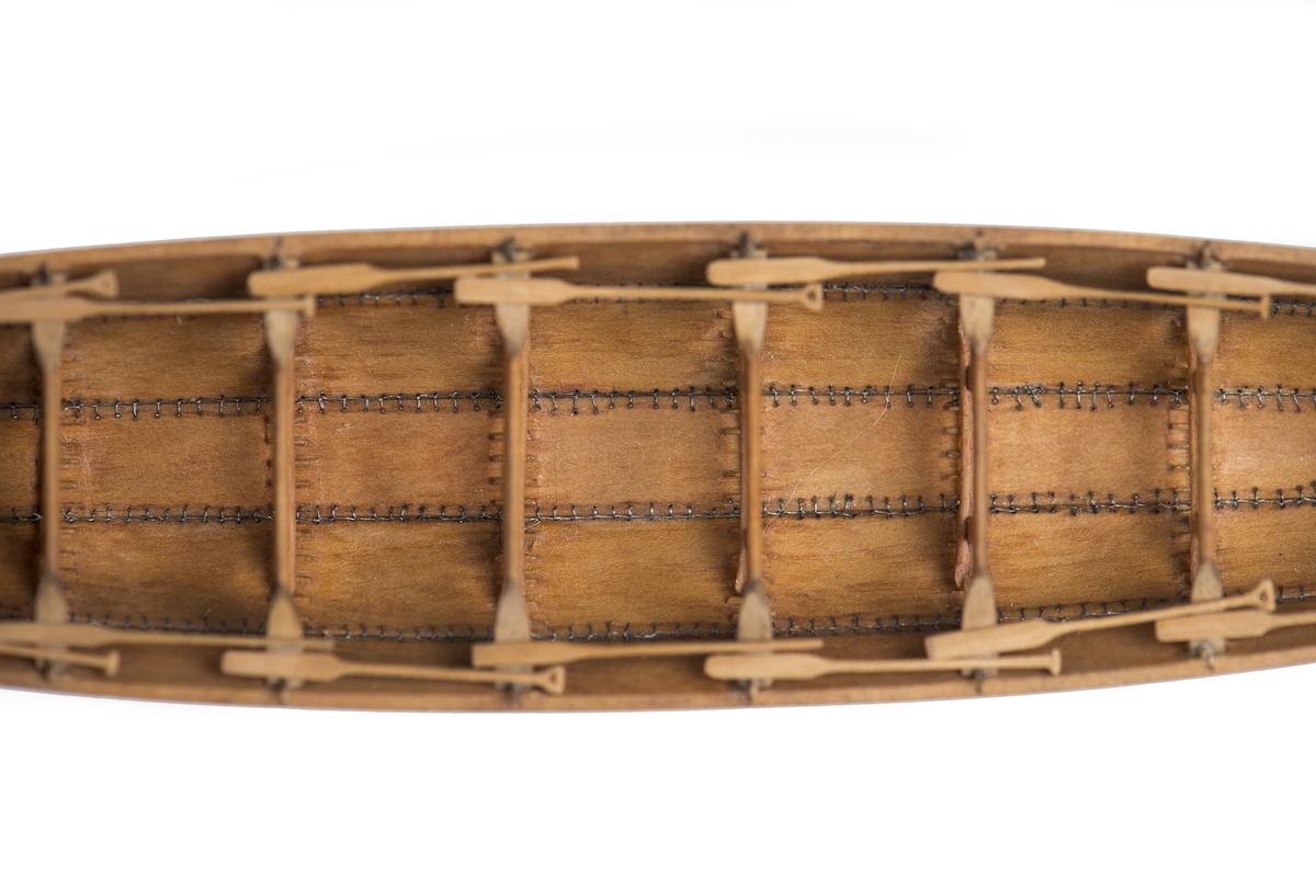 Modell av oxelträ, rekonstruktion av Hjortspringsbåten, utgrävd 1921, daterad till 200-300 f.Kr. Klamparna på borden utsparade i samma stycke. Spanten av piasava. Sammansyningen av borden, kölplankan, stävarna och spanten gjord med svartoxiderad suturtråd. Träet svagt betsat.
Lösa detaljer: 20 st paddlar, 1 st.styråra, 1 st. öskar.