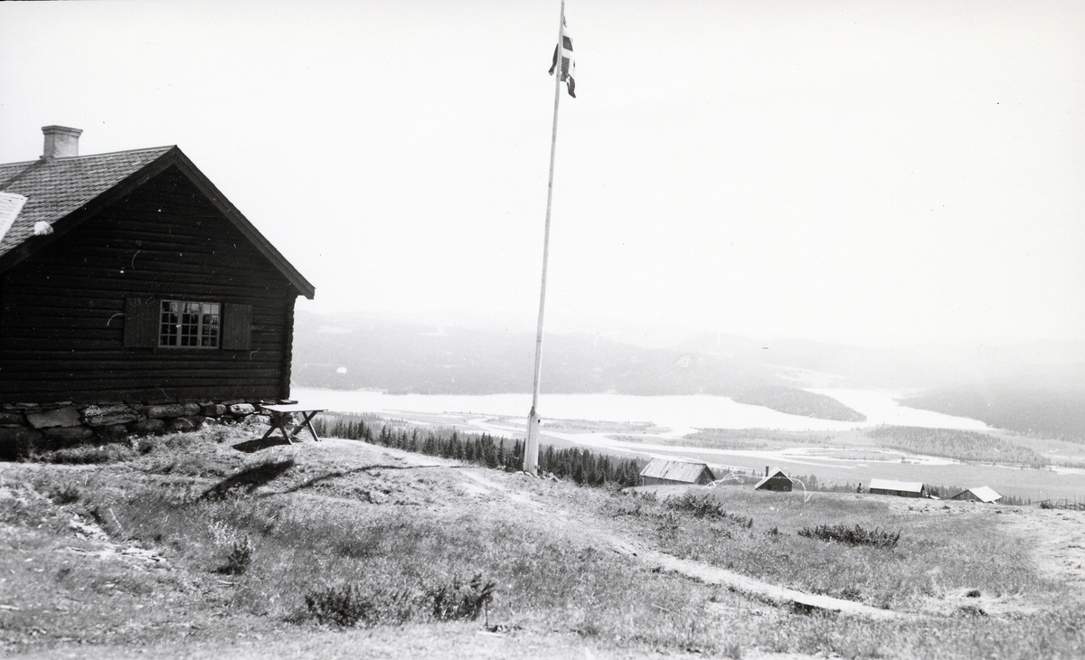 Tømmerhus ved flaggstang, med utsikt over stølslandskap og innsjø