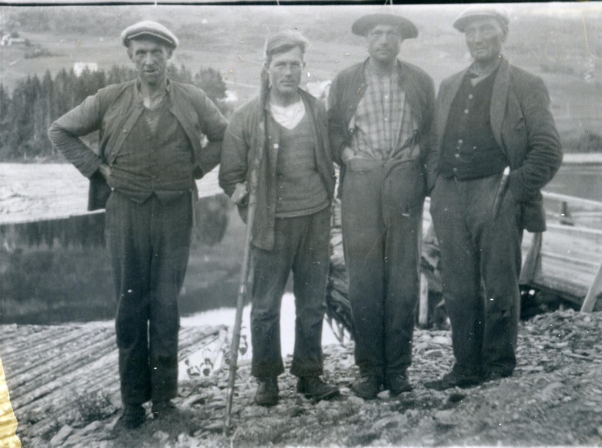 Fire menn oppstilt i forbindelse med tømmerfløting, en holder en båtshake som ble brukt til å dra til seg og styre tømmeret med. Mann nr 2 fra høyre er muligens Jon Tuv fra Røn i Vestre Slidre