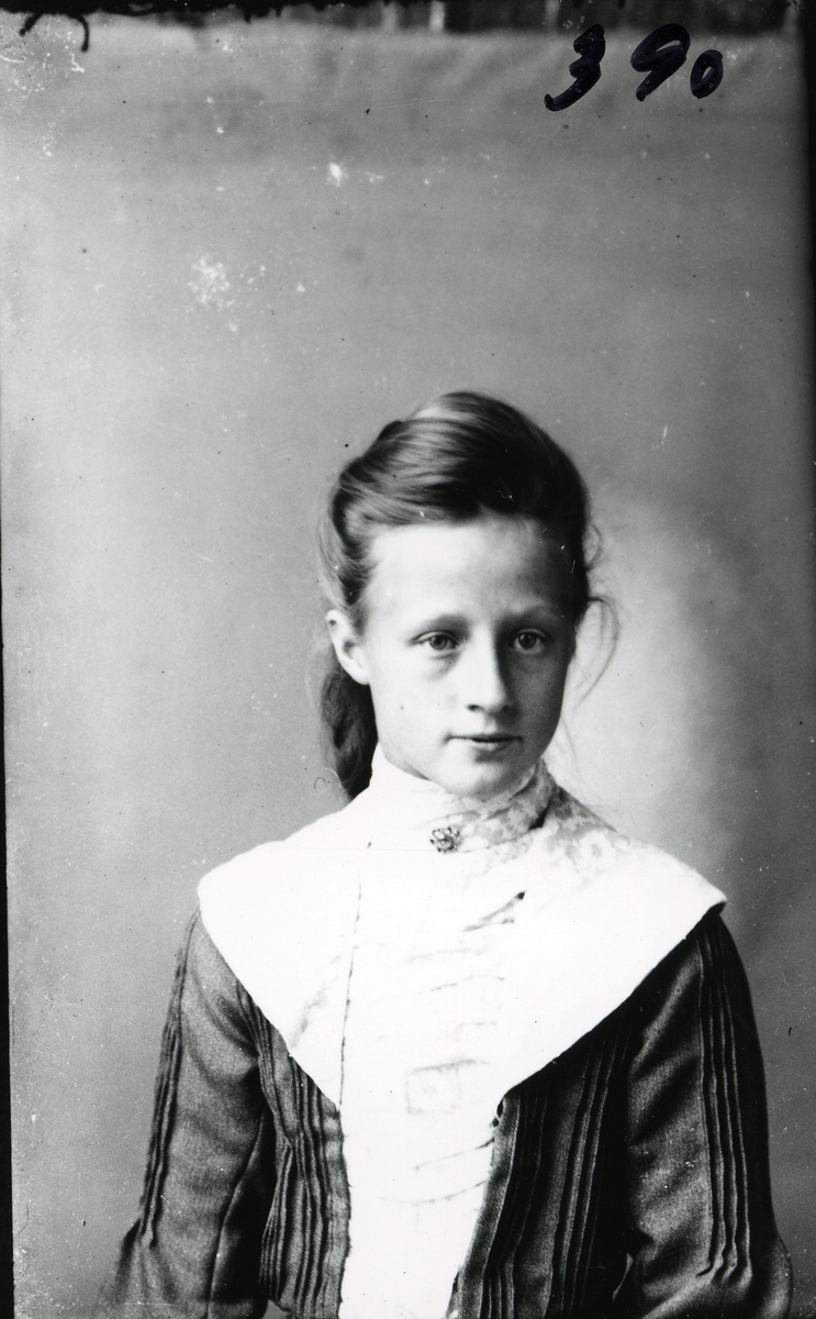 Portrett i halvfigur av ung kvinne. Mørk kjole med hvit krage. Halsnål.