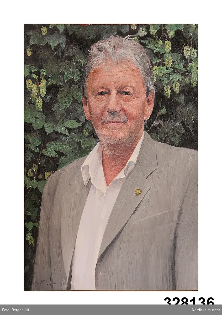 Porträtt föreställande Lars Löfgren klädd i grå kavaj och vit skjorta uppknäppt i halsen. I knapphålet sitter Nordiska museets märke med NM. Bakgrund av ymnig grön humle.
/2003-11-19 Sigrid Eklund