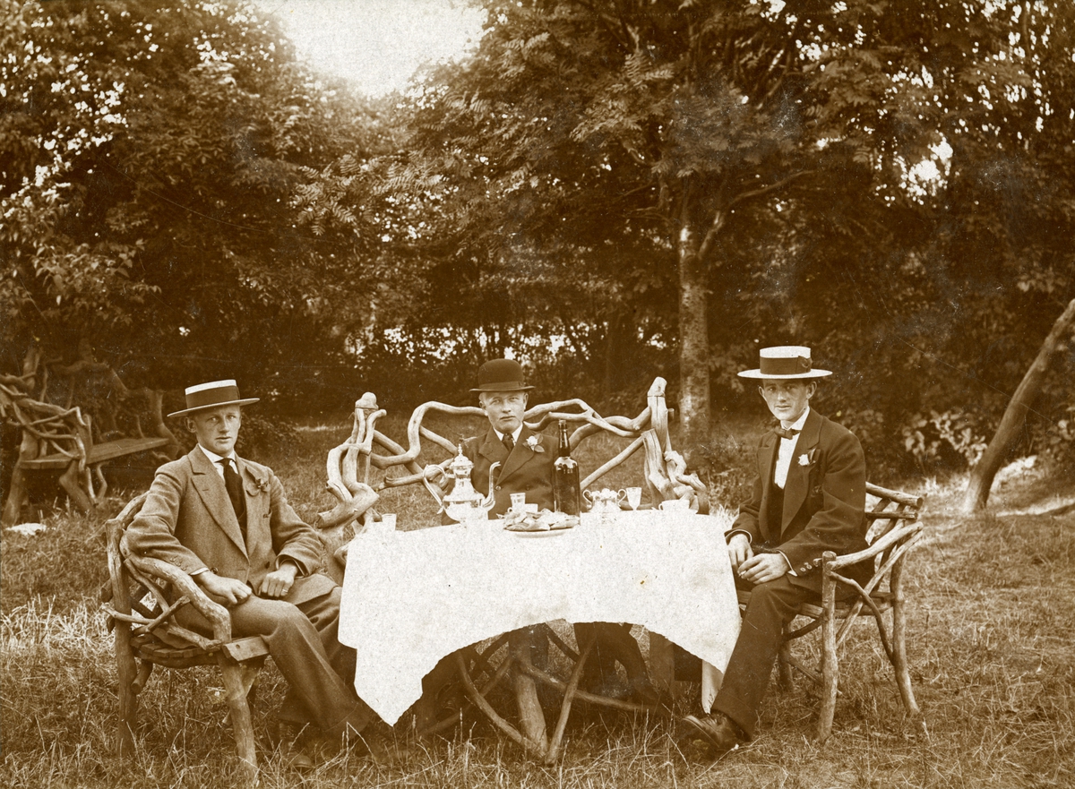 Civilklädda herrar sitter och fikar i en trädgård.