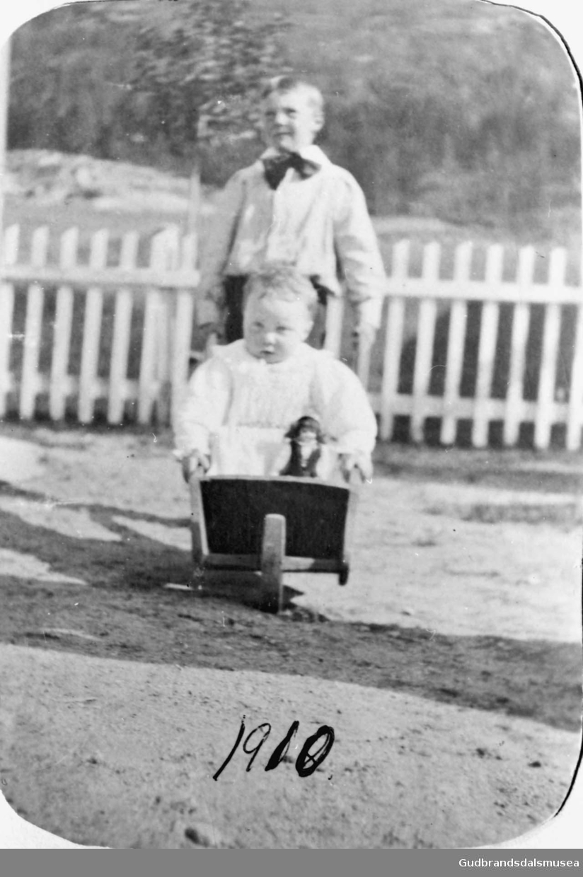 To barn leker med trillebåre ute i hagen/gårdsplassen? på Øygarden?, gjerde i bakgrunnen. 