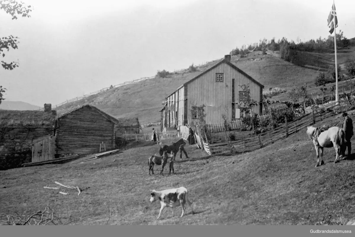 Gårdsplassen på Uppistugu- og Nistugu Kvam, fem personer ikke registrert, det er kyr og hester på gården, fjell og trær i bakgrunnen.
