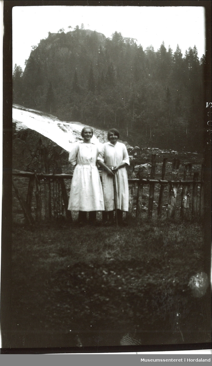 To kvinner som står med ett gjerde i hverdagsklær. Foss og fjell i bakgrunnen. Høyre kvinne holder på en stokk.