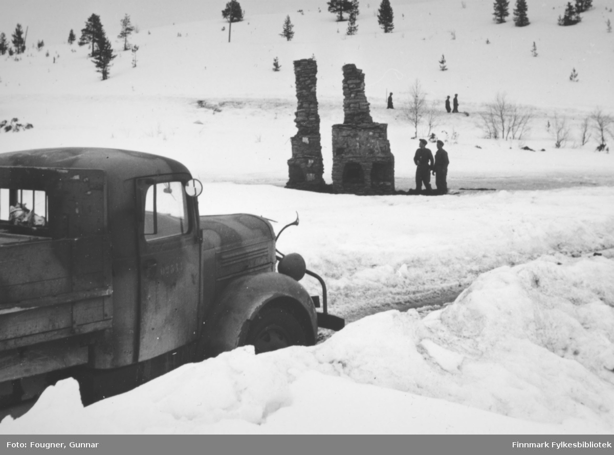 Fra Karasjok. En militær lastebil (Volvo "rundnese" kom på markedet i 1939, produsert til 1953/54, også i militære versjoner), malt i kamuflasjefarger kommer kjørende fra venstre. To soldater iakttar et par skor- steier som står igjen etter bombing.
