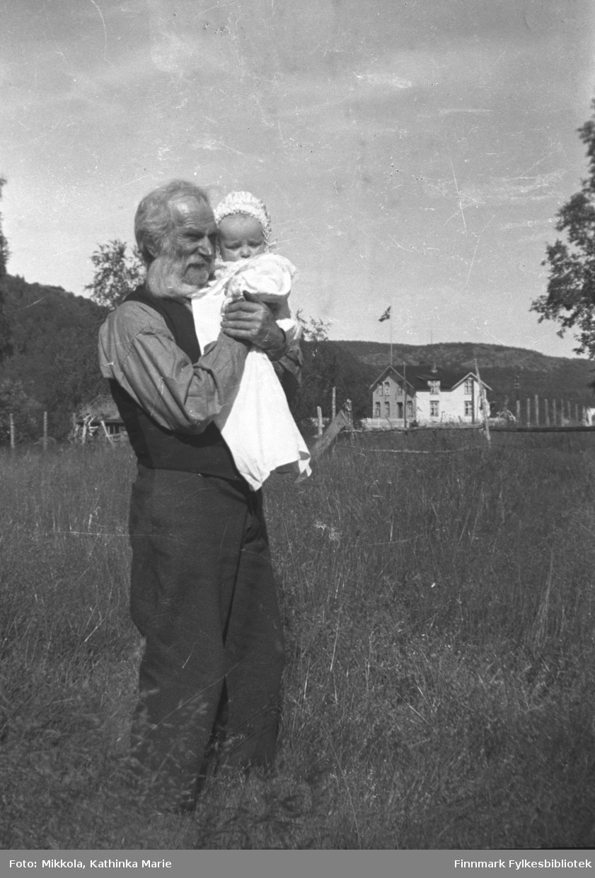 Mikkel Mikkola d.y. med et spedbarn, Aino Mikkola på dåpsdagen i 1937. I bakgrunnen John Haaheims hus. Bildet er tatt på eiendommen Nordmannseng i Neiden. Bildet er tatt ved samme anledning som 05007-388.