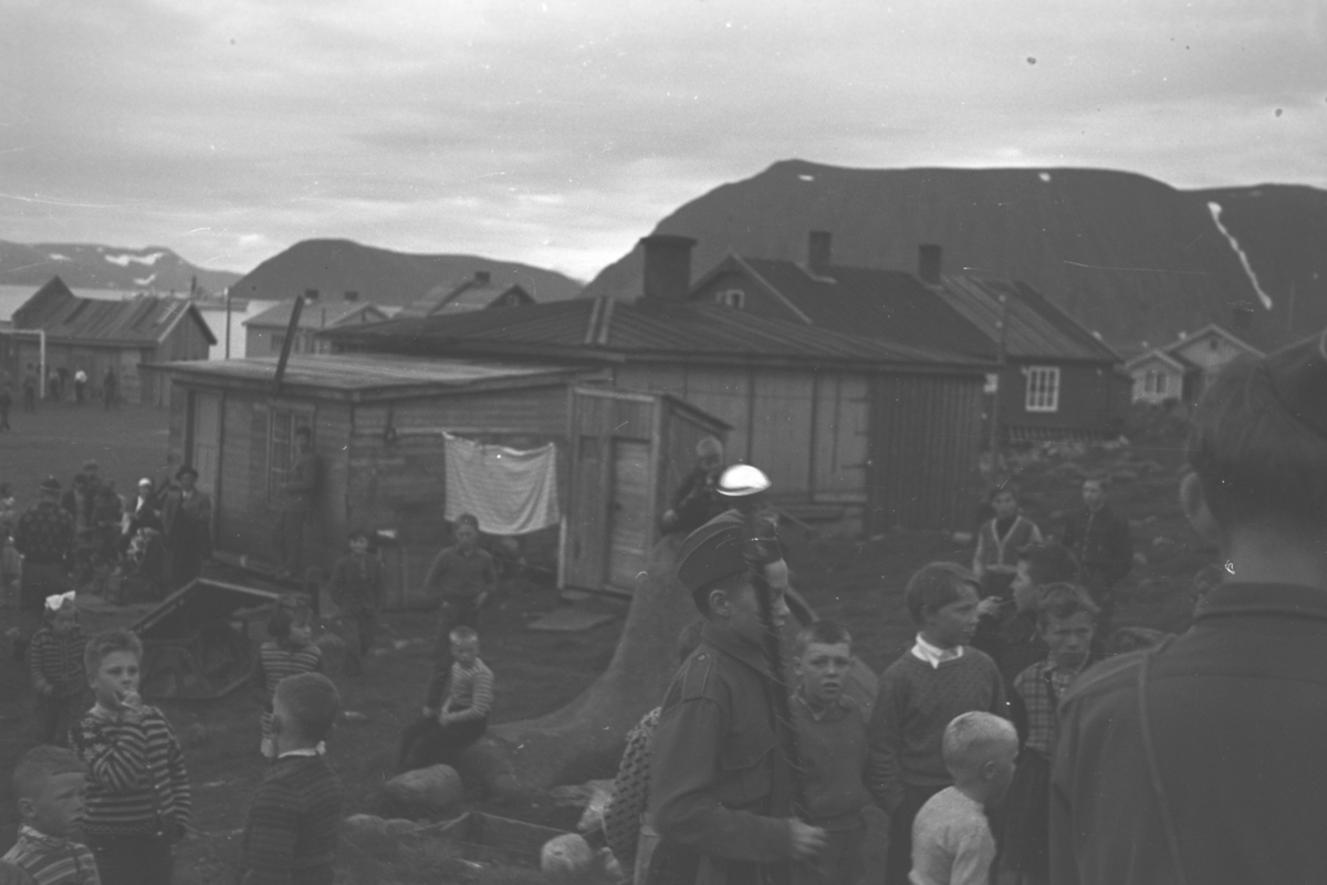 En gruppe barn er samlet på en slette foran noen hus og brakker. Sted og anledning er ukjent, men det kan være en korps- eller speidersamling.