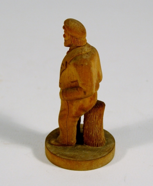 Figur av mann i dress og lue som lener seg mot en stubbe.