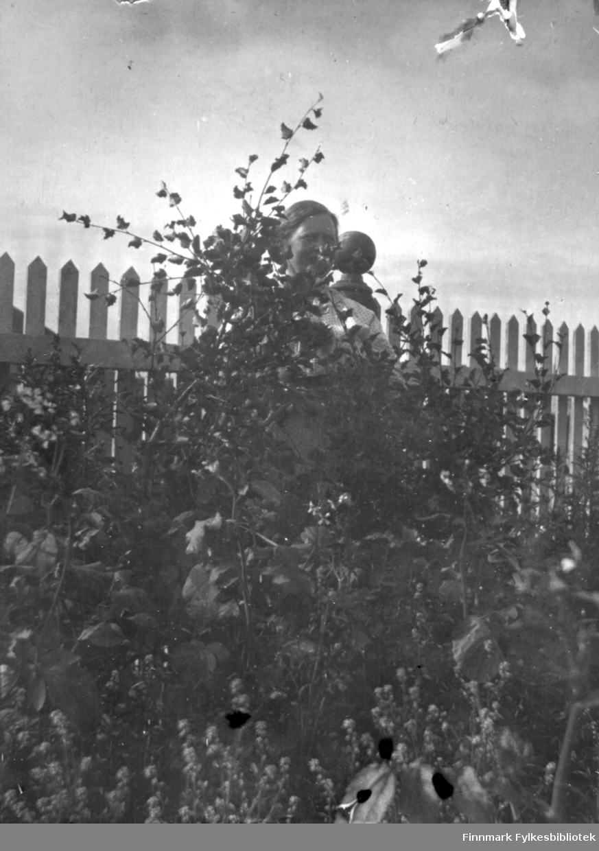 En dame i lys bluse fotografert i hagen til Arthur og Kirsten Buck på Hasvik. Det er ganske mye planter i hagen. Bak gjerdet til venstre på bildet ses såvidt en fjellside som går nedover. Et stakitt-gjerde  står i bakgrunnen. En hvitt gjerdestolpe med dreiemønster og kule på toppen vises ved siden av hodet hennes.