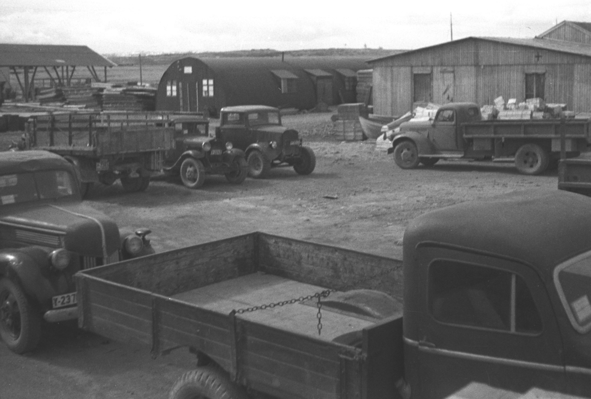 Gjenreisningstid. Flere lastebiler parkert på et område med brakker rundt. Stedet er ukjent, men kan være fra Ørtangen i Vadsø. Lastebil i forgrunnen med registreringsnr. Y-237 (og  trolig 4. siffer skjult) er en tysk- eller franskbygd Ford årsmodell 1941 eller nyere. Nærmest av lastebilene i bakgrunnen, med fronten frem, kan være en Ford AA 1930-31. Ved siden av kan være en Ford Thames WOT (=War Office/Krigsministeriet), bakerst i full lengde en Chevrolet 1946-47 med typisk militær lasteplan. Kan også være bygd 1941 eller senere for det amerikanske Forsvaret. Bak de to Fordene Nissenhytte - halvrund engelsk militærbrakke i metall.