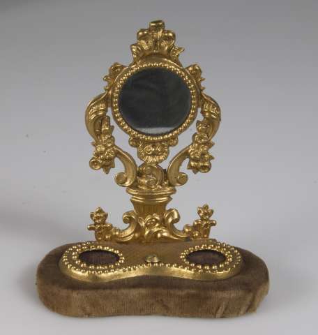 Toalettspegel av gulmetall stående på träskiva klädd med brun sammet. Spegeln hör till byrå UM08385e_01. 