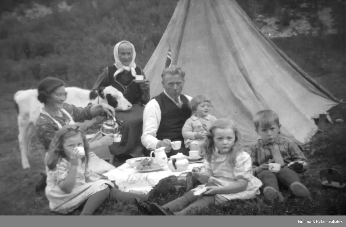 En sommerdag ved gården Fjellstad, øverst i Tverrelvdalen i Alta. På bildet ser vi familien Johansen som sitter på bakken, utenfor et telt. Det er dekket med duk på gresset. Oppå står det kopper, fat med kaker, en mugge, og en kanne. "Moster" Karen Kristine Thomassen (f. 1879) som har et hvitt tørkle på hodet, sitter på en stol bakerst. Moster Karen Kristine var søster til Elen Marie (f.1852) som var gift med Christen Arnesen. Etterslekten fikk derfor navnet Kristensen. Karen Thomine sitter her sammen med sine barn og ektemann. Pappa Andreas Julius er kledd i hvit skjorte, slips, og vest. Han har Anna Elisabeth 2 år på fanget. Jentene Karen Kristine 6 år, og Ragnhild Marie 4 år, er kledd i hvite kjoler. Karen har hårbånd. Gutten som sitter til høyre er et søskenbarn. Han heter Jens A. Kristensen, og er 6 år. Han er kledd i en stripete skjorte. Bak pappa Andreas Julius ser man en del av det norske flagget. Familien Kristensen er en stor familie spredt på stedene Elvebakken, Nerskogen, Transfarelv og Tverrdalen. 