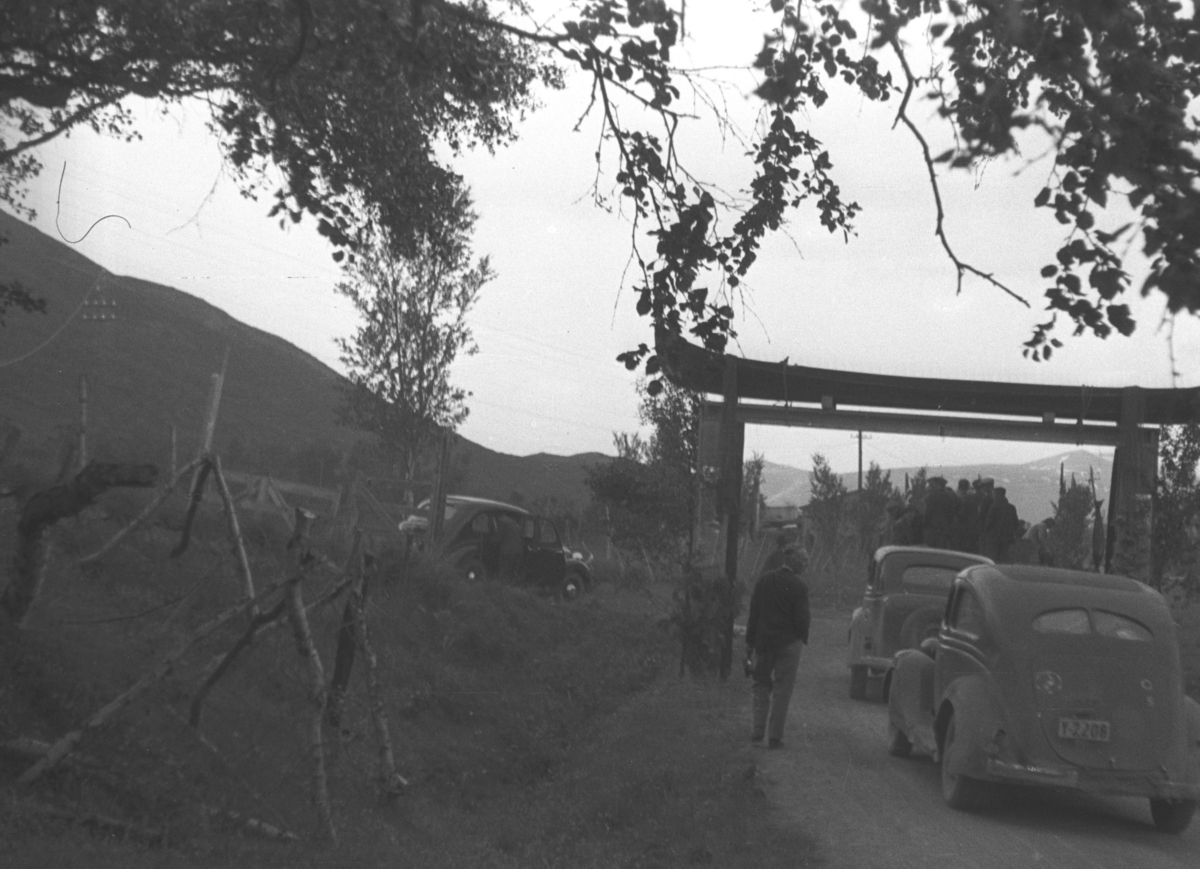 To biler kjører under en slags portal på en skogsvei og et par personer går ved siden av. Personer og sted er ukjent.