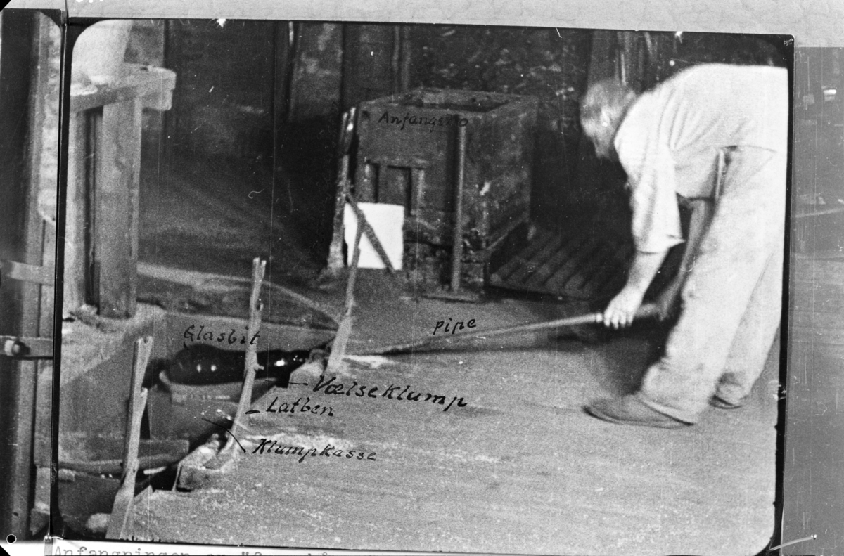Reprofoto av fotografi fra Drammens Glassverk. Motivet viser et interiør fra en produksjonshall. Bildet har påskrift med forklarende tekst. Beskrevet i motivet erglasbit, latben, klumpkasse, vælseklump og pipe.