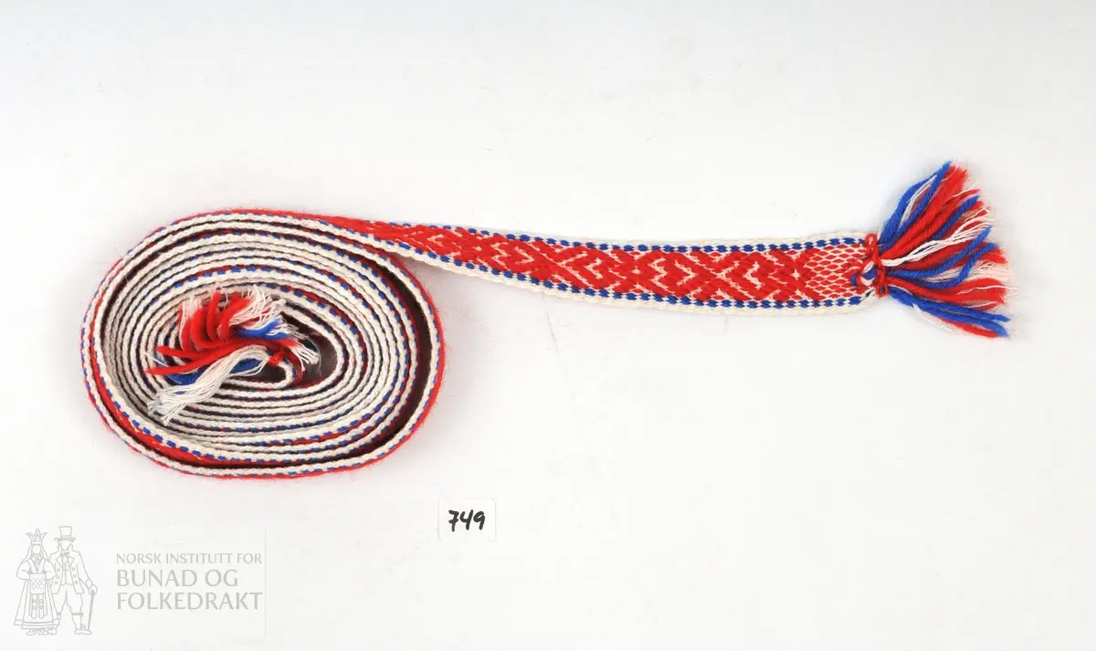 Band vove av kvit bomull og raud og blå ull. Innslag av kvit bomull. J.B.S innvove i eine enden, 1910 i den andre.