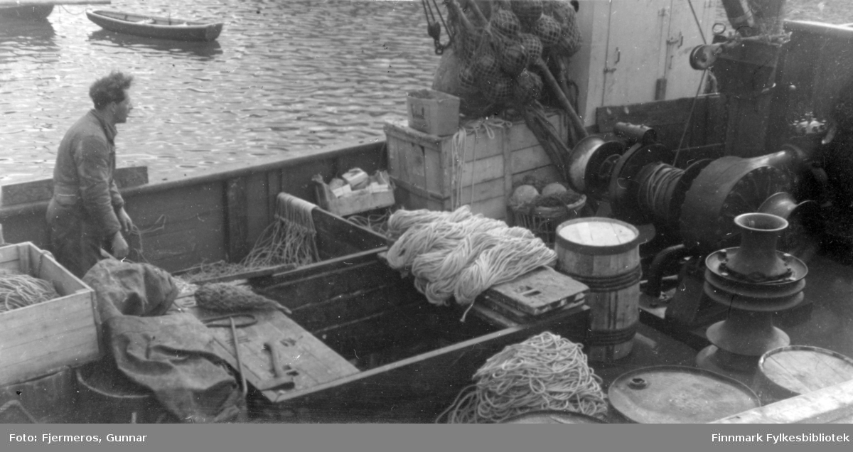 En mann på dekket til en skøyte som akkurat har tatt ombord utstyr tili å fiske/fange Håkjerring. Person er ukjent, men båten kan være "Varfjell" som muligens ligger ved kai i Kjøllefjord.