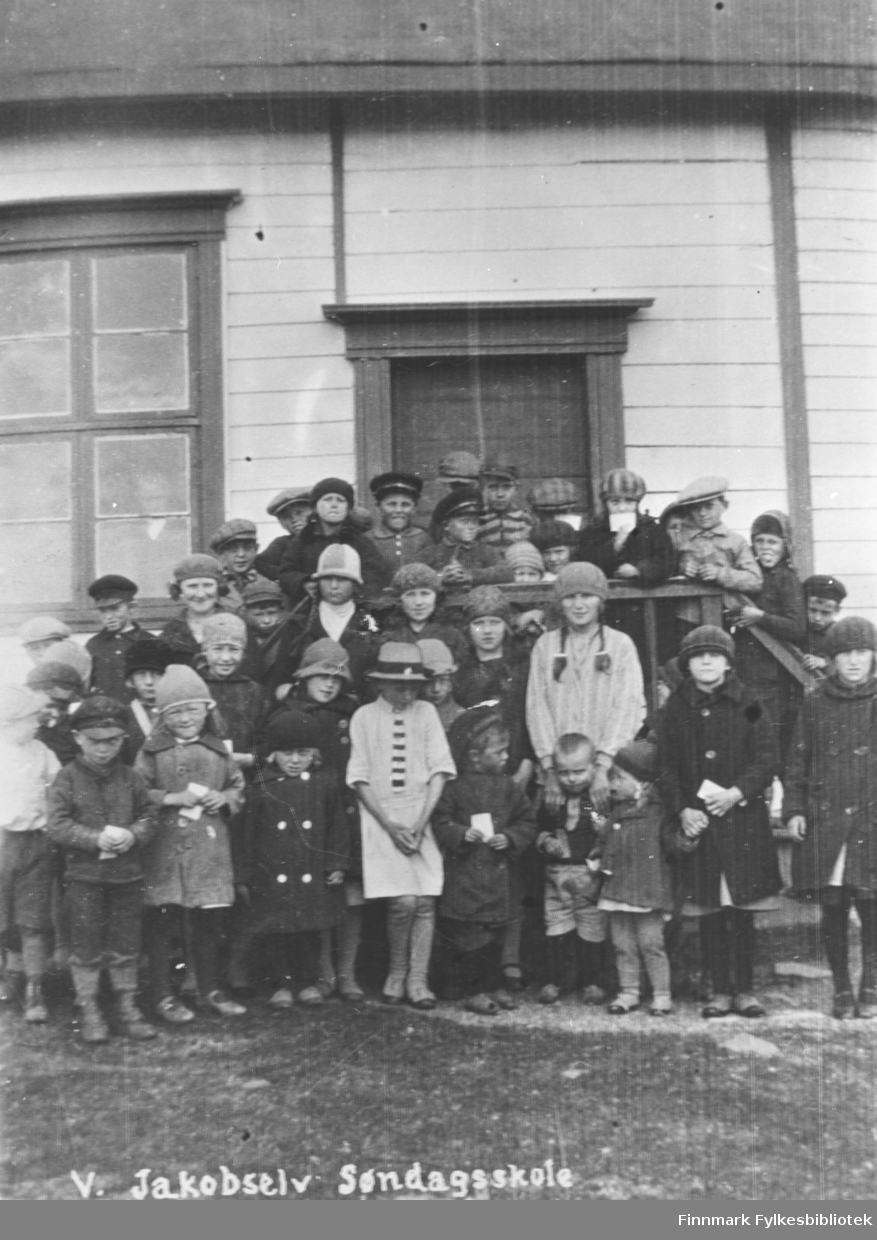 41 barn fotografert ved søndagsskolen i Vestre Jakobselv. En del av ungene står på trappa og de andre foran dem. Ungene er mellon 3-15 år gamle. Alle har hatter eller luer på seg og har en hvit lapp i handen. Bildet muligens tatt før 1930.