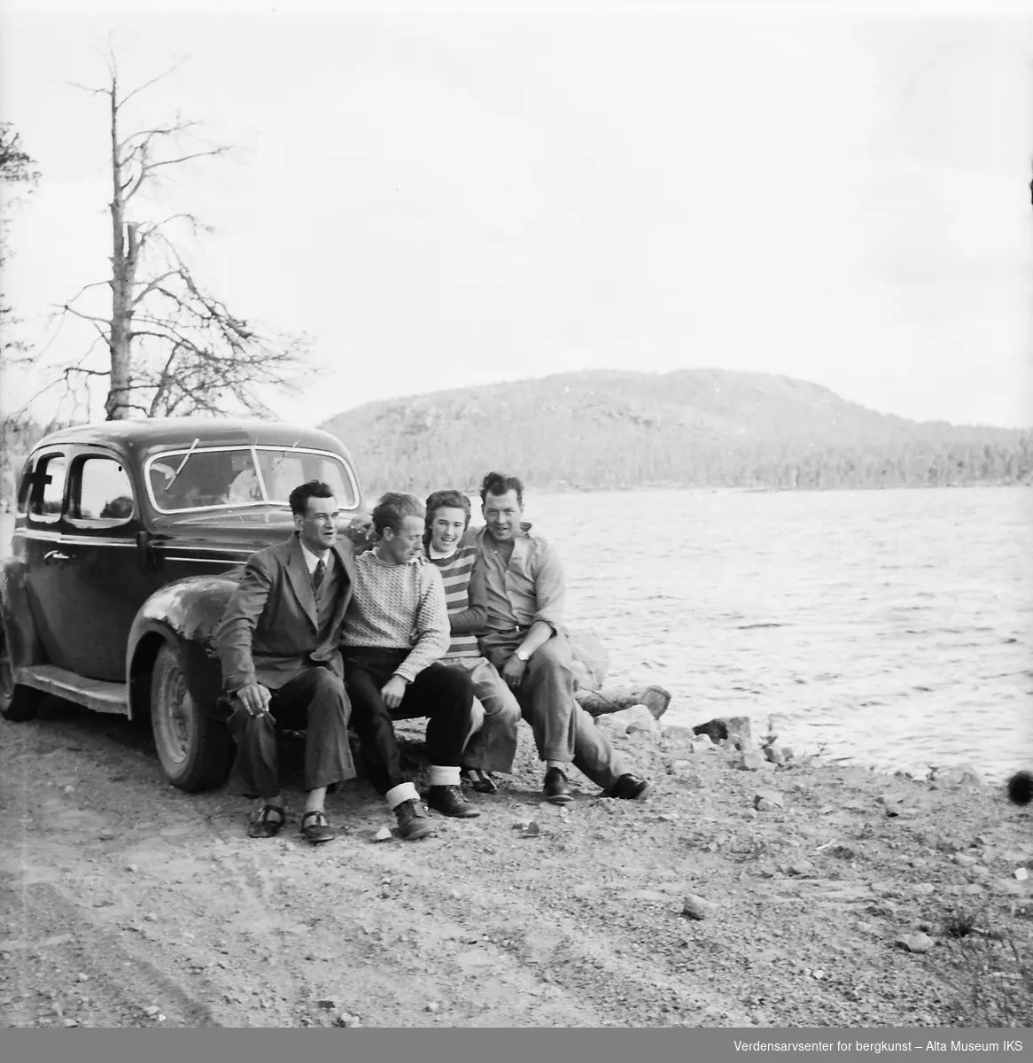 Fire reisende sitter på støtfangeren til bilen, med en innsjø i bakgrunnen. Bildet er fra en Finlandstur.