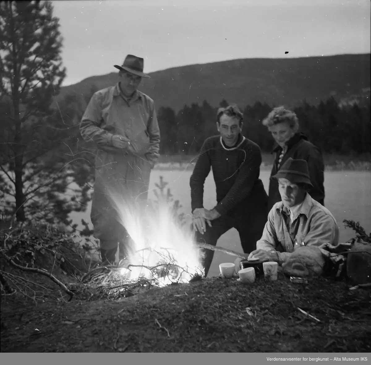 Fire personer sitter rundt et bål på elvebredden og drikker kaffe.
