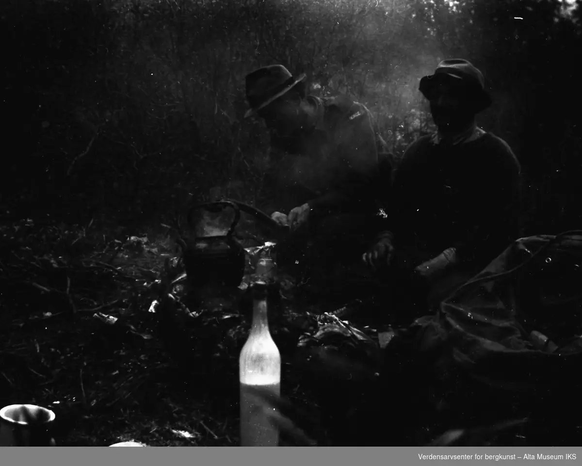 To menn iført hatt sitter ved et bål i skogen. De har med seg segger, og en høy flaske står foran bålet. En kaffekjele står på bålet.