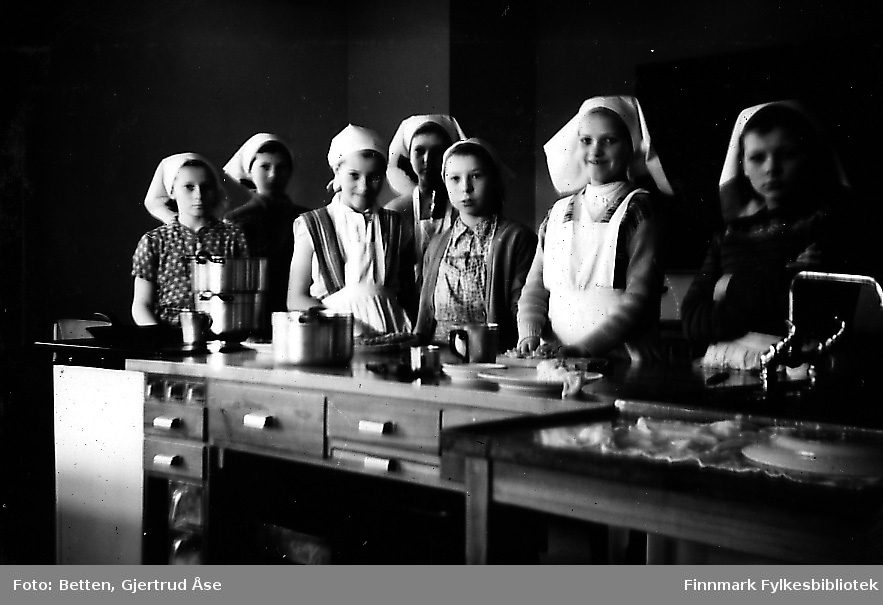 Jenter fra Smalfjord på kjøkkenopplæring i Seida skole og internat. Syv jenter kledd i forklær og tørkler står inne i skolekjøkkenet ved kjøkkenbenken. Jente nr 2 fra høyre heter Ragnfrid Johannessen (gift Mathisen).  Astri Dalsbø skal også være en av disse jentene.