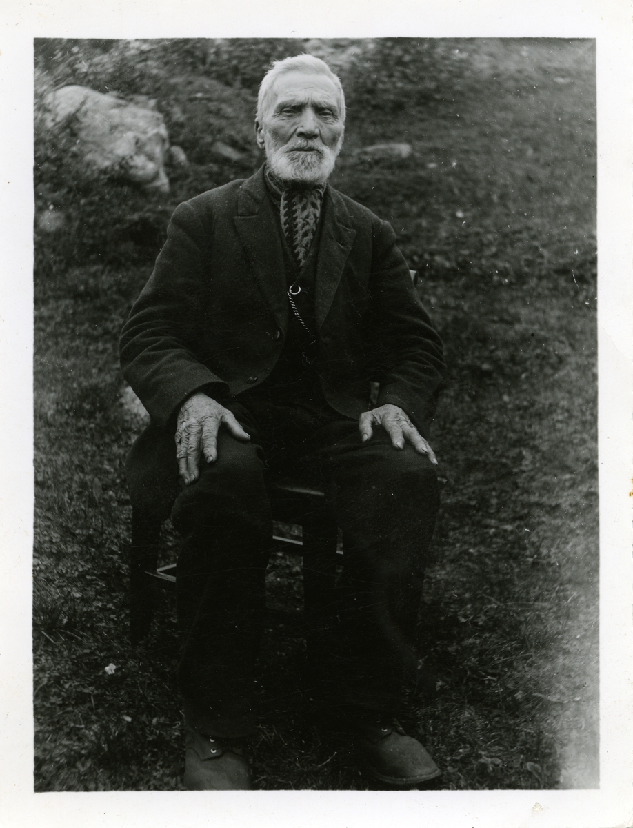 Portrett av mann sittende på en stol i en hage. Mannen er iført dress med skjerf i halsen.