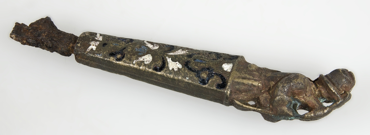 Knivskaft av brons med emaljinläggningar i vitt och mörkblått. Skaftet är längst upp format som en man som spelar säckpipa. Av knivbladet finns endast rester kvar.
Märkt "EC.603."