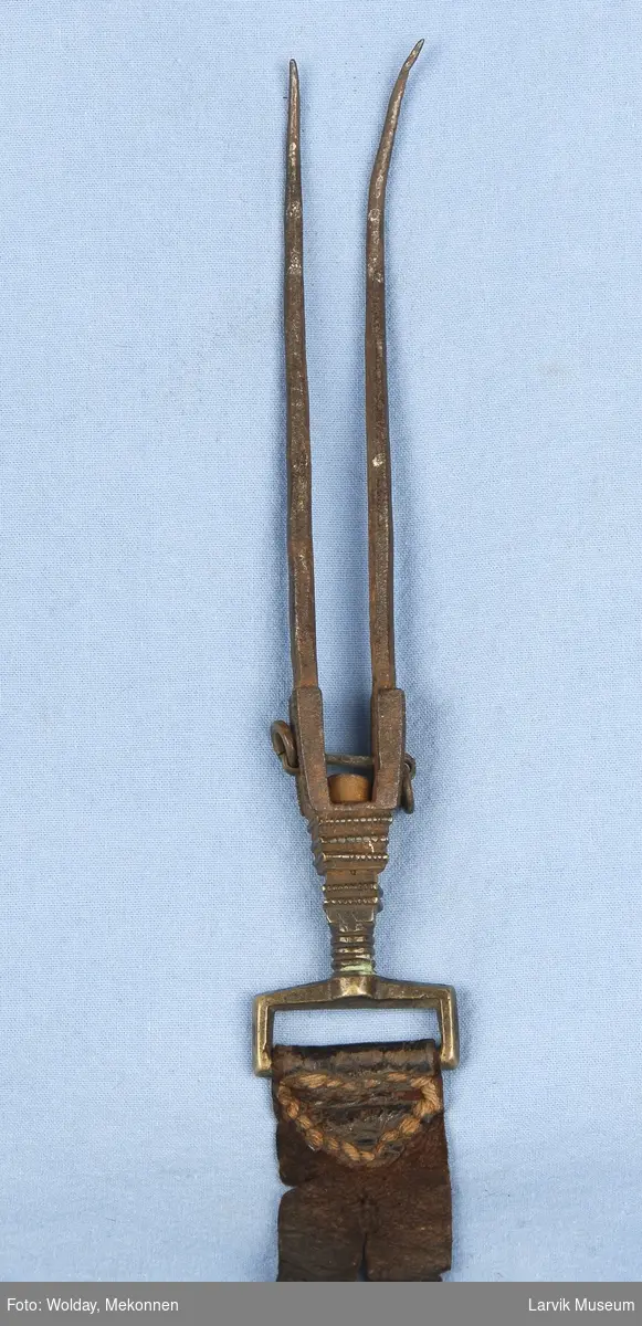 Form: To lange "gaffeltenner"som er noe bøyd i tuppen, festet i en skinnrem. Den er satt sammen slik at gaflene kan snurre.

