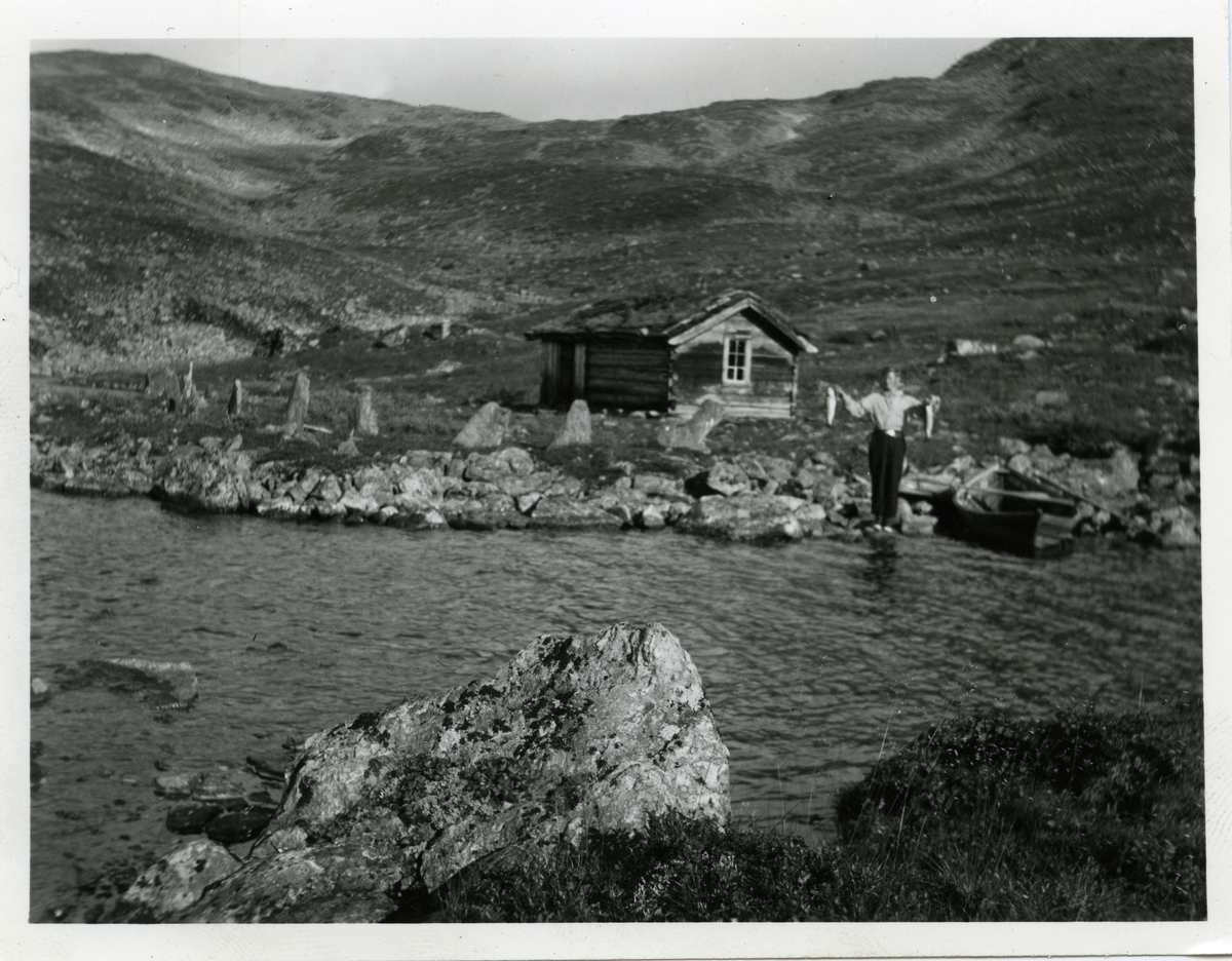 En mann avbildet foran en hytte ved et vann, og  med to fisker i hendene.