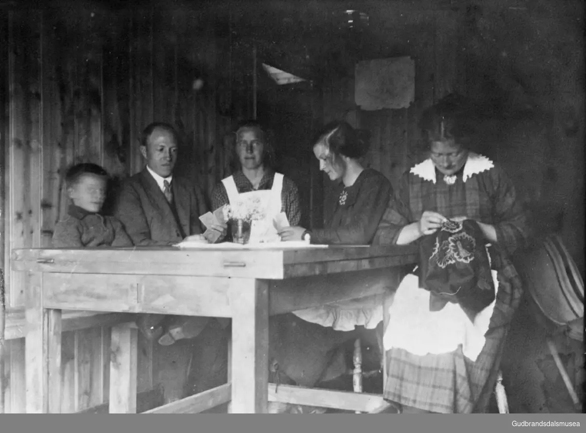 Fem personer ved bord, tre kvinner hvorav den ene sitter og broderer, de andre holder på med kortspill, på kjøkkenet i Nordsletten?

