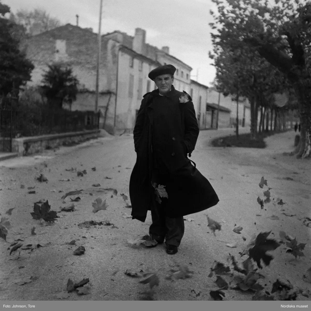 Man i rock och basker på gata täckt av kringblåsande höstlöv.