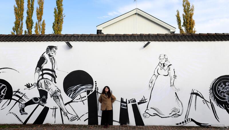 Lucia Aragon foran sinn veggmaleriet «Drømmer over grenser». Det viser en mann med redningsvest som møter en kvinne i bunad. Innvandring møter utvandring.