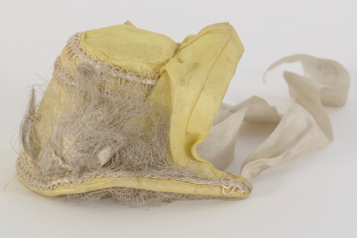 Dukkekyse i (oppr) gul silke med pyntebånd. Avstivet og pyntet med tyllkant og bånd. Foret med silke. Knytebånd i silke.