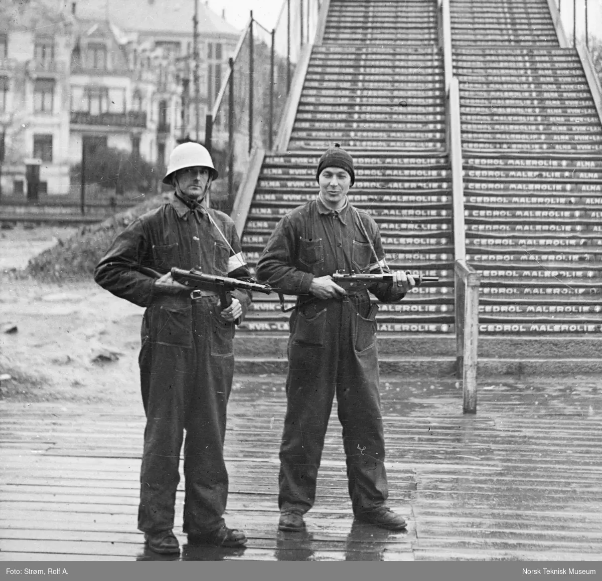 To menn med geværer, en med hjelm, den andre med lue, begge i arbeidstøy foran en trapp, trolig til en tribune, Oslo, mai 1945,