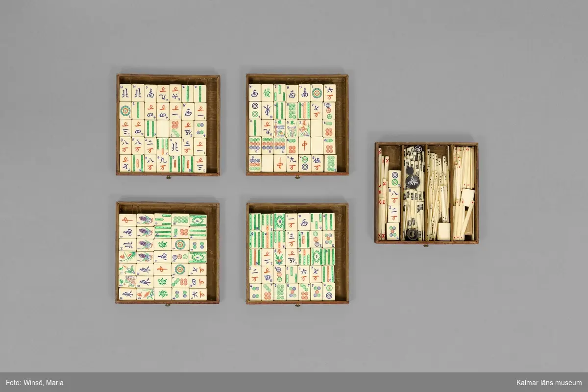 KLM 46455:1-2. Spel. Sällskapsspel, Mahjongspel (:1), bestående av: Fyrkantig låda, klädd i brunt läder, med två handtag i mässing på ovansidan. Inuti fem stycken utdragbara lådor med mässingsknoppar, innehållande spelpjäser av elfenben, med utskuren dekor, målade i grönt rött och blått. Tillhörande folder med spelregler på franska (:2). Text framsida folder: "LE Mah-Jongg et comment on y joue Règle complète du véritable Jeu Chinois. Text ovansida: RUDA MAH-JONGG. Text botten: KIRBY, BEARD & CO, PARIS L.