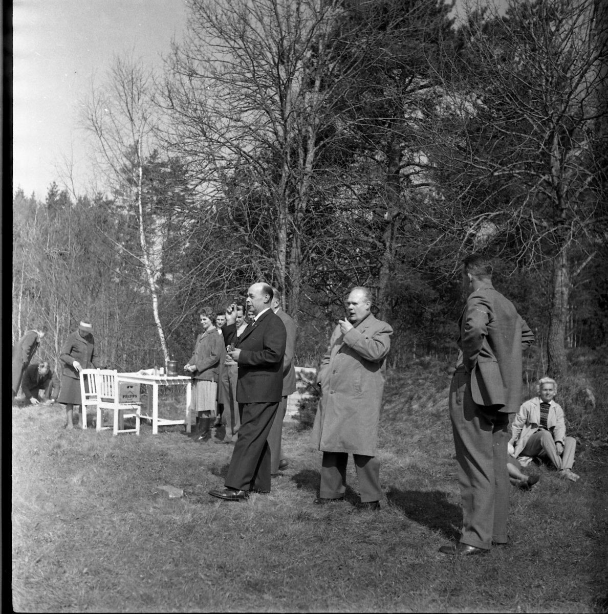Män och kvinnor ute i det gröna. En grupp med trädgårdsmöbler till vänster. I bildens centrum en man i mörk kostym som sannolikt spelar Dart. Intill står en man pipa i munnen, det är Holger Andersson.
