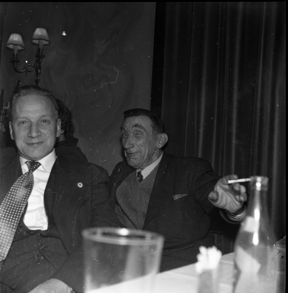 Två män, sannolikt på en restaurang. Ett glas och flaska i klarglas i förgrunden. Mannen närmast i bild med prickig slips heter Nils Johansson. Bakom honom sitter Gustav Larsson med ett plirigt leende och cigarett i handen.