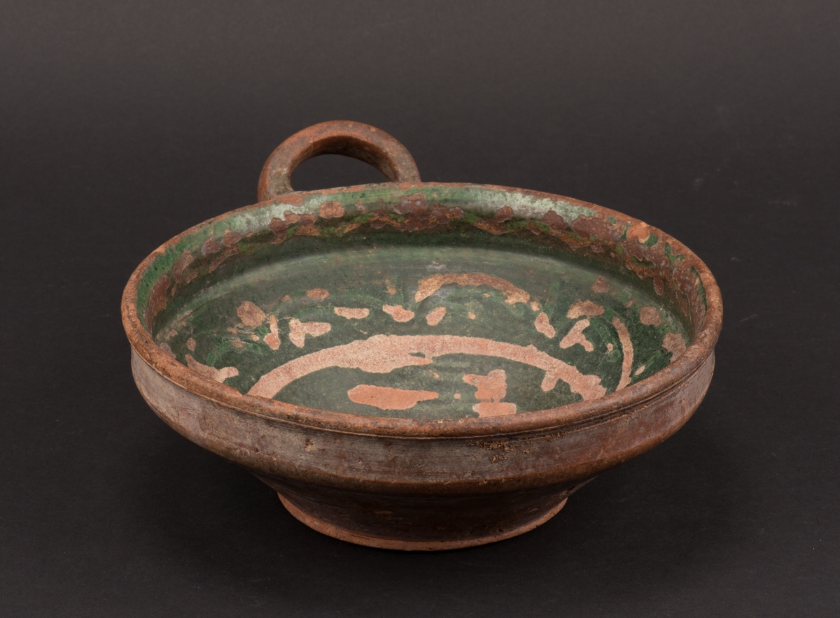 Rund, drejad skål av bränd lera. Konisk form med rak övre kant där skålen är försedd med ett öra. Grön glasering inuti skålen, med ränder och vågformat mönster samt årtalet 1830.