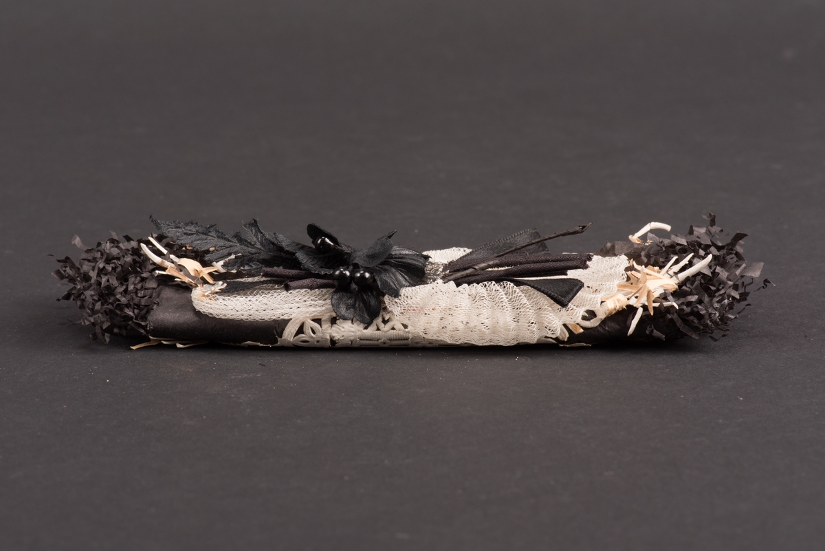Platt begravningskaramell inlagd i svart silkespapper med klippta och krusade ändar. Omlindad med ett vitt perforerat papper. På ovansidan ligger en blomma med en rosett i sidenband på en bit vitt stärkt tyll.