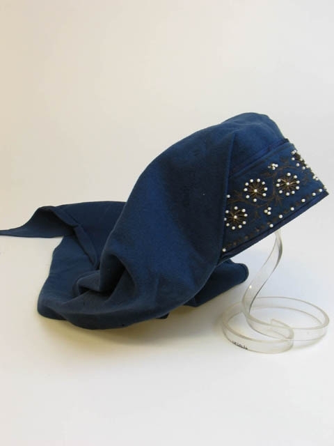 Huvudkläde av blått ylletyg; på hjässan bård broderad med vita pärlor och guldtråd uppsatt på mörkblått sidentyg.
L. Från snibb till snibb 160 cm.
Frk. Siri Malmros, 1958
