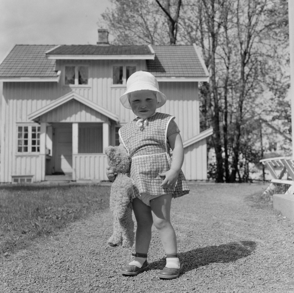 Tangen, Gjerdrum. Anne 2 år med kjole og solhatt på tunet med bamsen