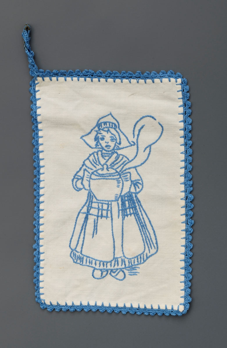 Pynteklut med påbrodert figur i blått garn på hvit bunn og blå heklet kant og heklet hempe i ene hjørnet. Firgurmotivet er en jente m. tradisjonell hollandsk påkledning. Nr. 2 av totalt 6 kluter.