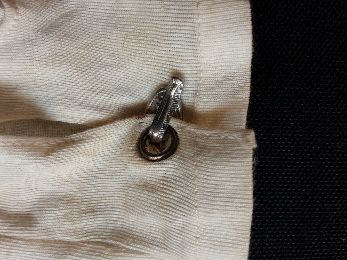 Hanske. Venstre. Hvit bomull, splitt på innsiden av håndleddet som lukkes med metallspenne. Tre maskinbroderte hvite striper på handbaken.