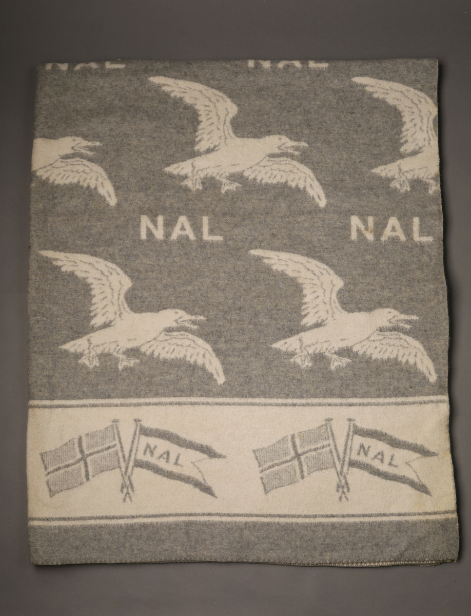 Ullpledd med måkemotiv i fargene naturhvitt og grått. Produsert for Den norske Amerikalinje. Felt nederst på hver side med norske flagg og NAL-flagglogo.