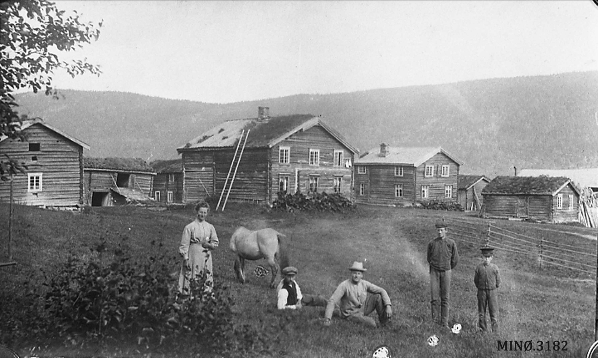 Kjernefamilie foran gardskompleks, arbeidsantrekk, hest. 