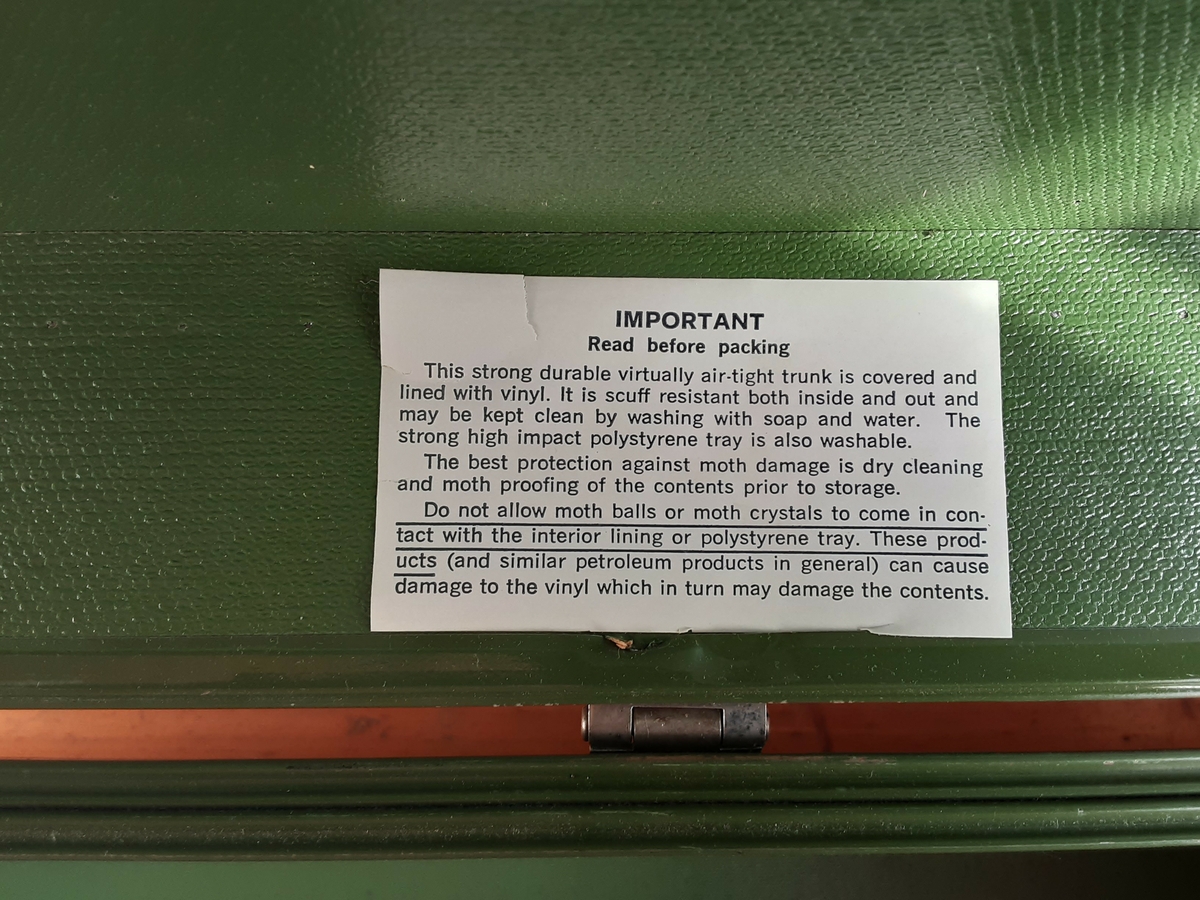 Koffert dekket med grønn vinyl, kantene forsterket med bånd og hjørnene med metallbeslag. Lærhåndtak på hver kortside.