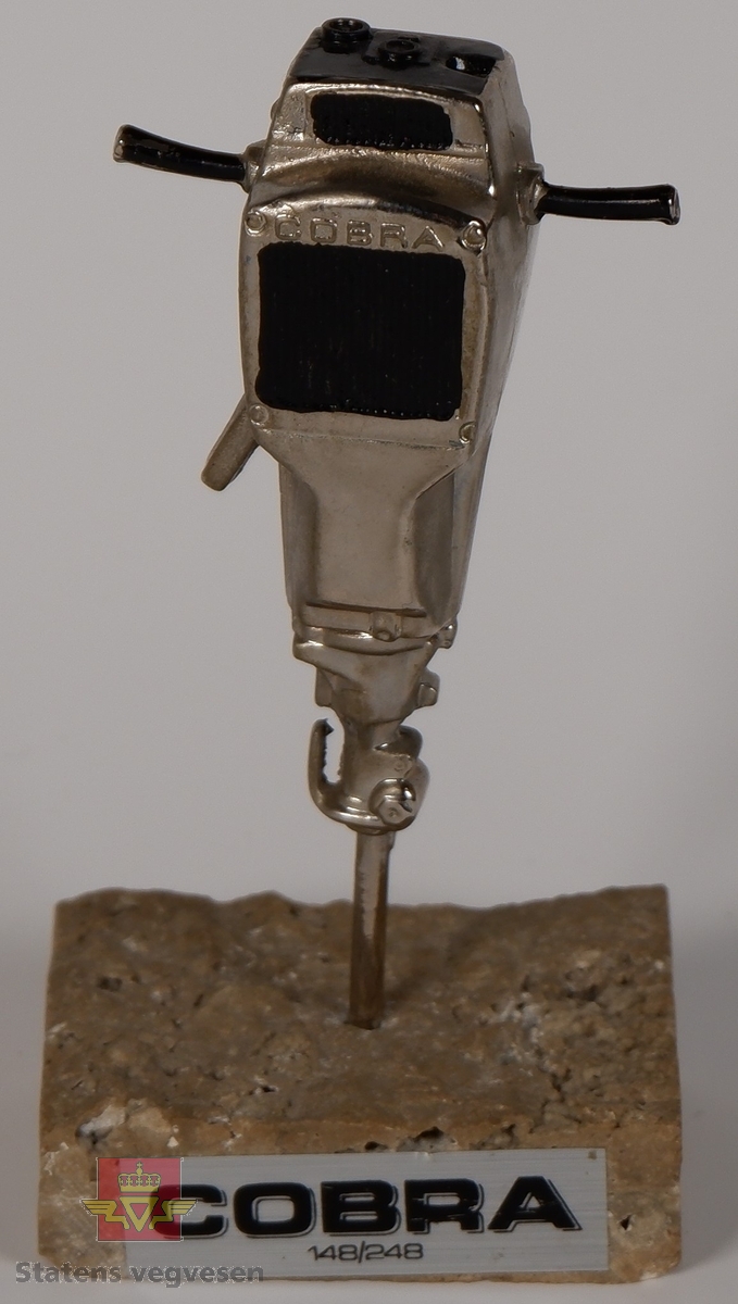 COBRA. Sølvfarget miniatyr boremaskin i metall som står med boret festet i en sokkel av stein. Boremaskinen har innskriften COBRA, og på steinsokkelen er det et klistremerke som har påskriften COBRA 148/248.